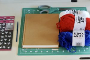 A picture of the materials needed for the article on How to make a tassel.  Materials included in the photo are yarn, a book, scissors, a crochet hook, a cutting board, and star stickers. 